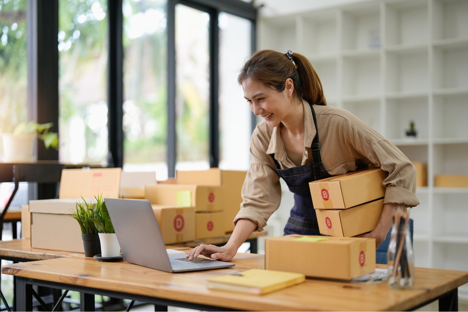 Small business owner with boxes in hand, working on laptop.